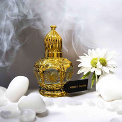Elegant gold bottle of White Oud attar surrounded by white stones and flowers, with soft smoke in the background.